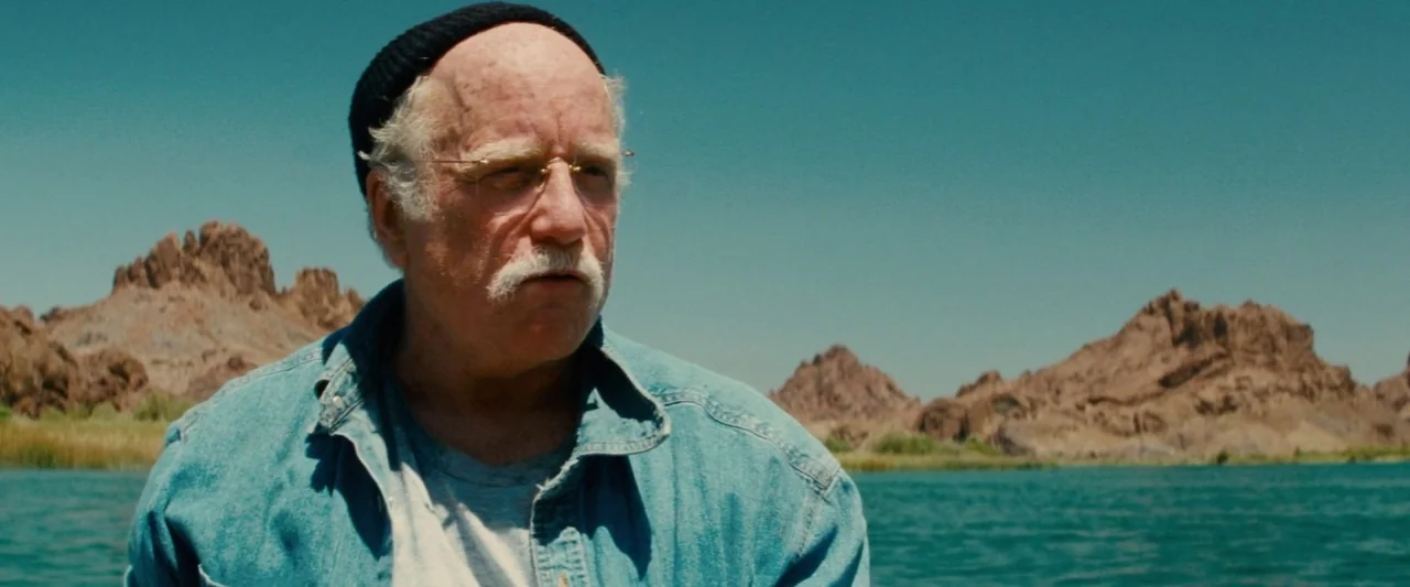 Piranha 3D (2010) movie still: medium — A sunny, desert landscape features a calm lake with rocky brown hills in distance. An older man in a blue…