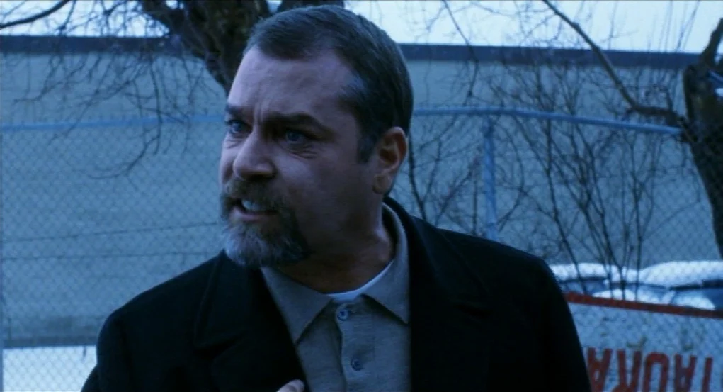 Narc (2002) movie still: medium — A man stands outdoors against a background of a chain-link fence and leafless trees. He wears a dark overcoat and…