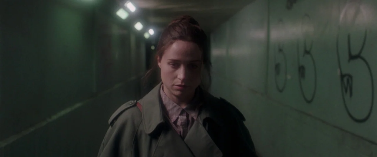 Censor (2021) movie still: medium — A narrow concrete pedestrian tunnel with green walls and black graffiti. A woman walks toward the camera through…