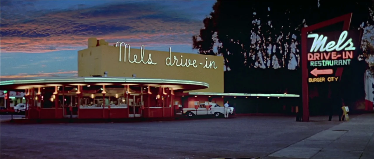 American Graffiti (1973) movie still: establishing — A 1950s style drive-in restaurant features a yellow building with neon cursive signage. The…