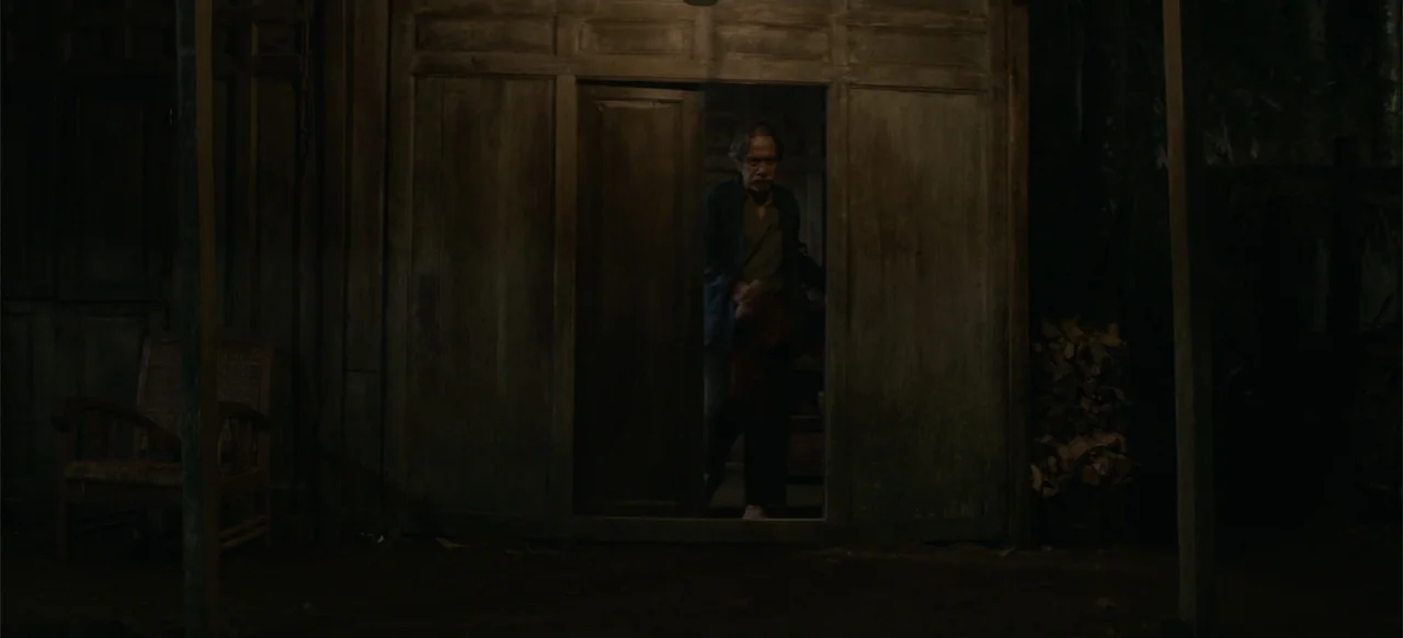 A Thousand Days (2023) movie still: medium — A dark wooden house exterior with a central doorway is visible at night. An older man with grey hair…