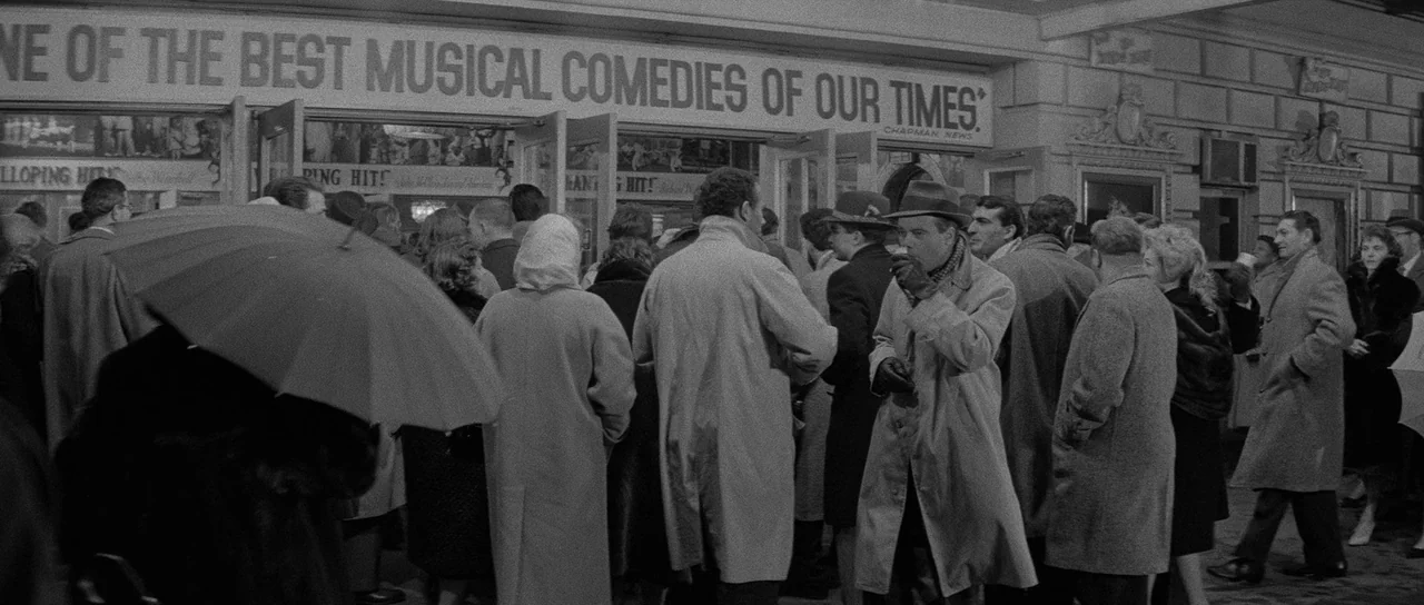 The Apartment (1960) movie still: wide — A crowd of people wearing heavy coats and hats waits outside a theater entrance. A large sign above the…