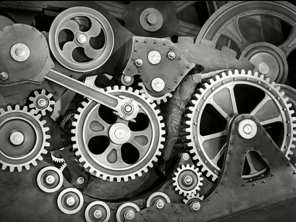 Modern Times (1936) movie still: wide — A complex network of large iron gears and industrial machinery dominates the frame. A single worker in…