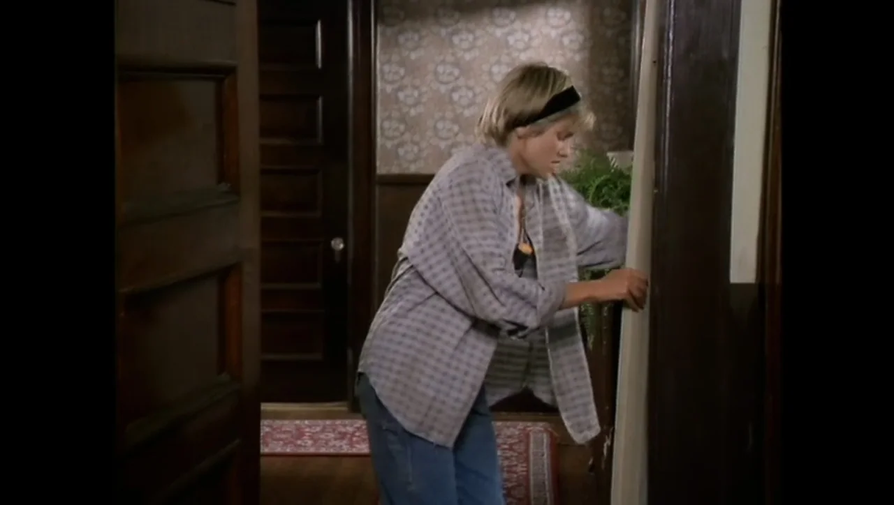 The Awakening (1995) movie still: medium — An interior hallway features patterned floral wallpaper and dark wood trim near a doorway. A woman stands…