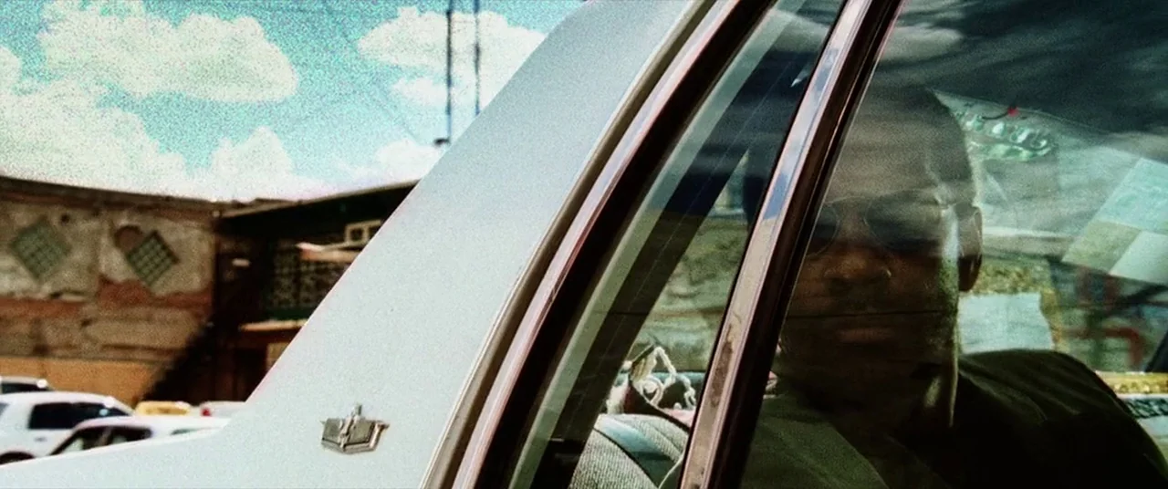 Man on Fire (2004) movie still: medium — A man sits inside a vehicle seen through a side window reflection. A parking lot with a brick building…