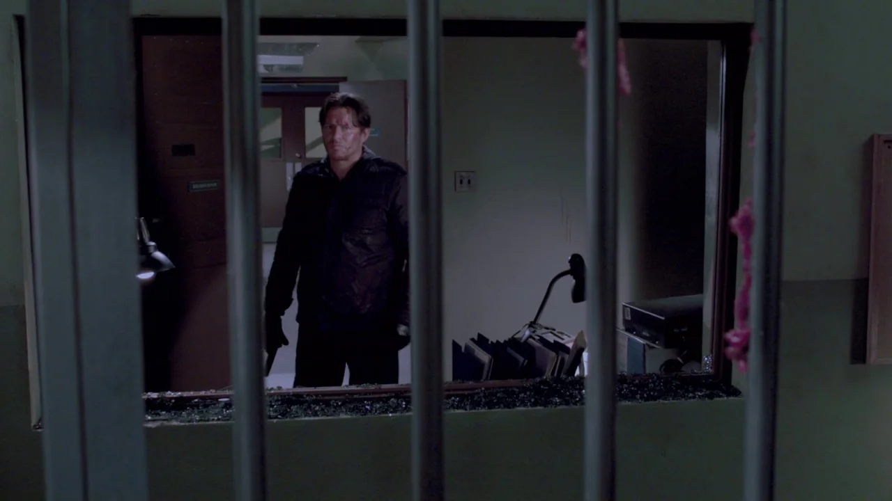 Saw VII: The Final Chapter (2010) movie still: medium — A dark corridor viewed through a set of metal bars with shattered glass debris. A man stands…