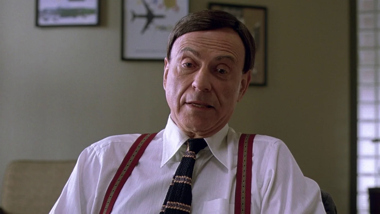 Eros (2004) movie still: medium — An office interior features plain beige walls decorated with framed prints of aircraft. An elderly man in a dress…