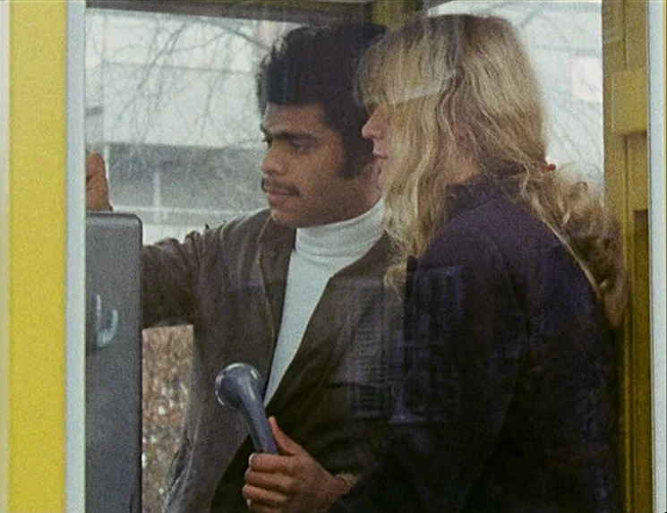Rio Das Mortes (1971) movie still: medium — A man and woman are inside a yellow public telephone booth outdoors. They stand side-by-side while the…
