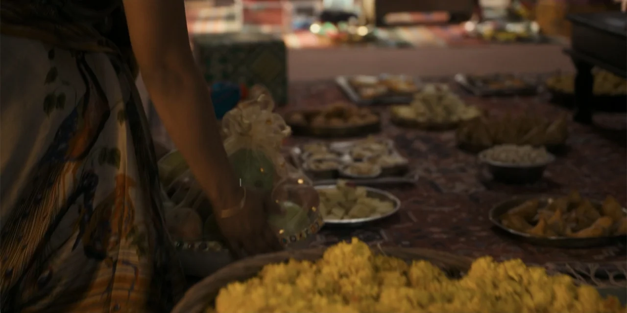 Dry Day (2023) movie still: medium — A low-angle view shows a table spread with various Indian snacks and dishes. A woman in a patterned garment…