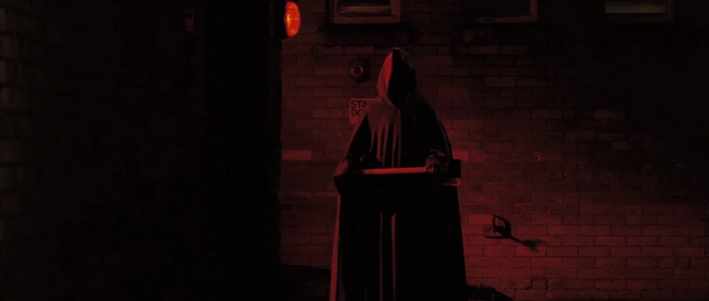 Hot Fuzz (2007) movie still: fullbody — A dark brick building exterior is cast in heavy shadow at night. A hooded figure stands perfectly still in…