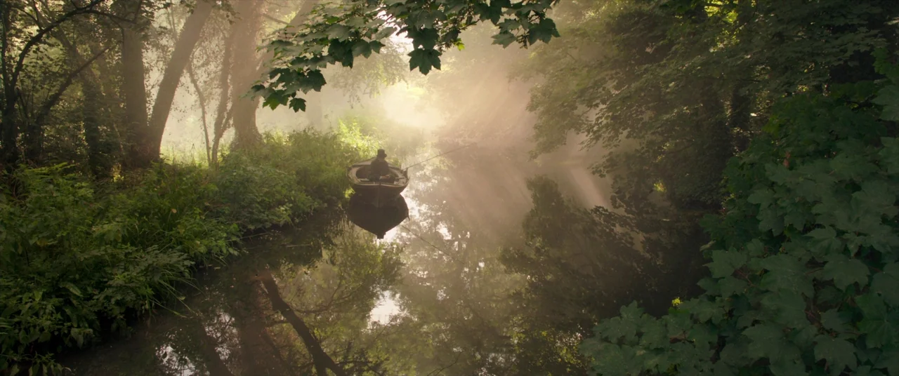 Mr. Turner (2014) movie still: wide — A quiet, narrow river winds through dense foliage under a thick morning fog. A lone person sits in a wooden…