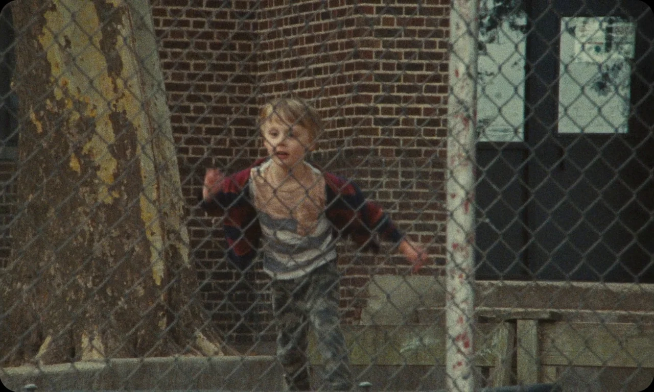 Vulcanizadora (2024) movie still: fullbody — A school playground with a red brick wall and large tree behind a fence. A young boy runs in the…