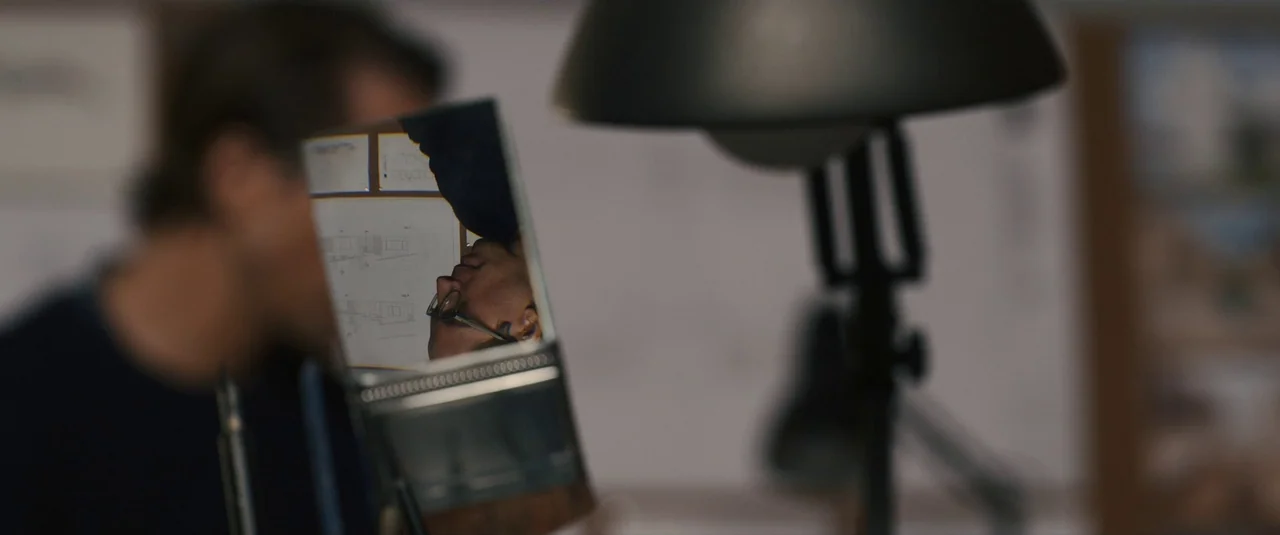 Intrusion (2021) movie still: closeup — A dark workspace features a desk lamp and architectural drawings visible in a mirror. The man's face appears…