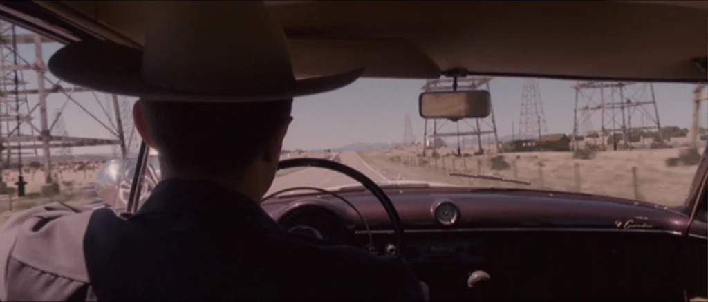 The Killer Inside Me (2010) movie still: medium — Inside a 1950s car looking out over the steering wheel at a desert road. The driver wears a cowboy…