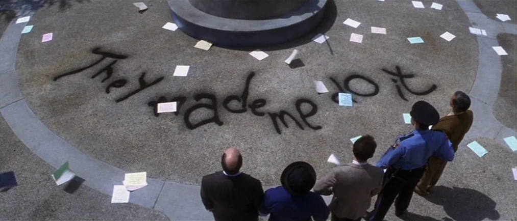 Donnie Darko movie still: wide — A group of men stand around a circular stone base in a paved plaza. Spray-painted black text reading 'They made me…