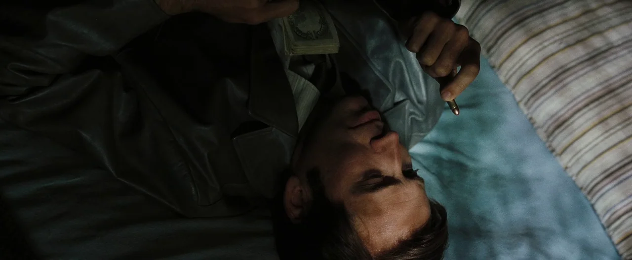 Lord of War (2005) movie still: closeup — A man lies on a bed with striped bedding in a dimly lit space. He holds a single brass bullet in one hand…