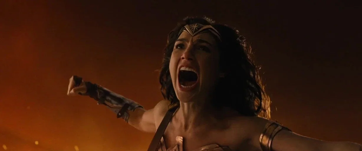 Wonder Woman (2017) movie still: closeup — A dark, smoky battlefield at night with warm embers floating in the air. A woman with a metallic tiara…