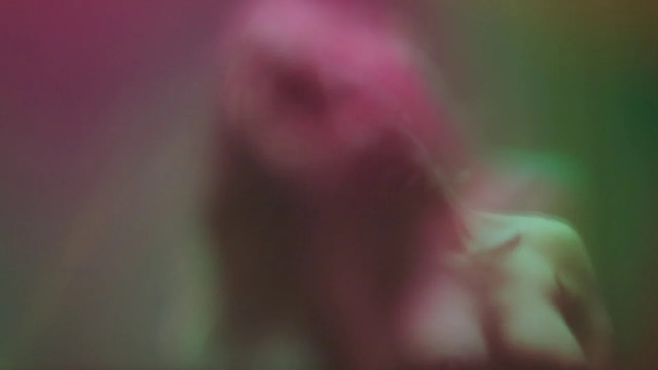 Infinity Pool (2023) movie still: closeup — A person is visible behind a frosted or wet glass pane. The figure is heavily blurred with pink and…