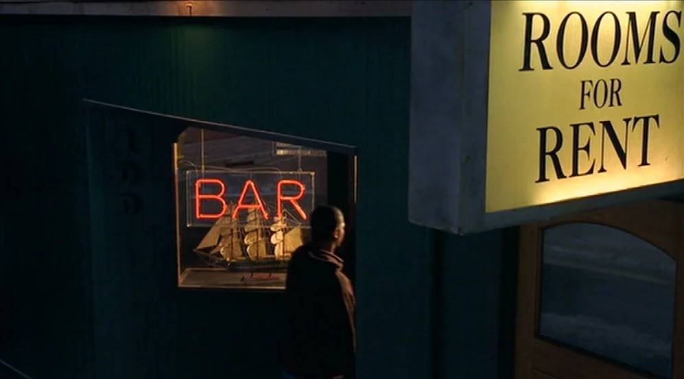 Limbo (1999) movie still: wide — A dark green exterior wall features a glowing red bar neon and a sailboat model. A man faces the window while a…