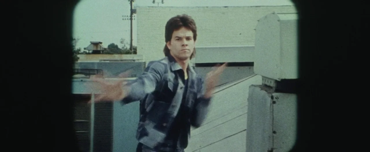 Boogie Nights movie still: medium — A flat rooftop of a commercial building with HVAC units and white walls. A young man with a mullet performs a…