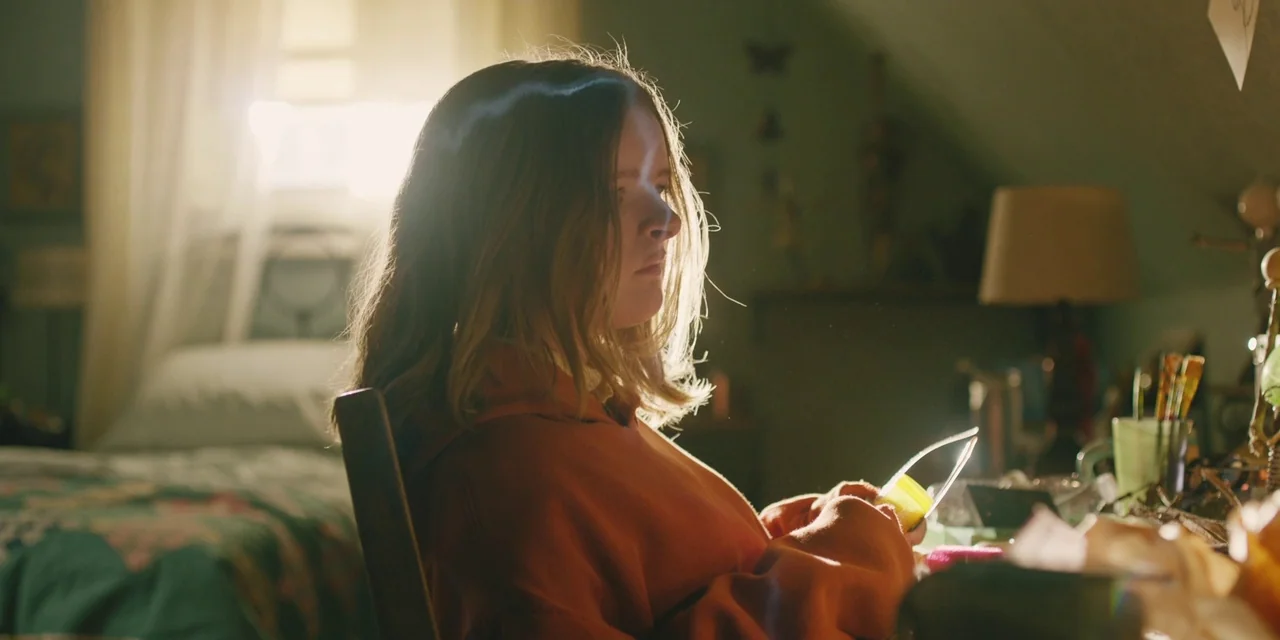 Hereditary (2018) movie still: medium — An attic bedroom with slanted walls contains a bed and a cluttered desk. A teenage girl sits at the desk,…