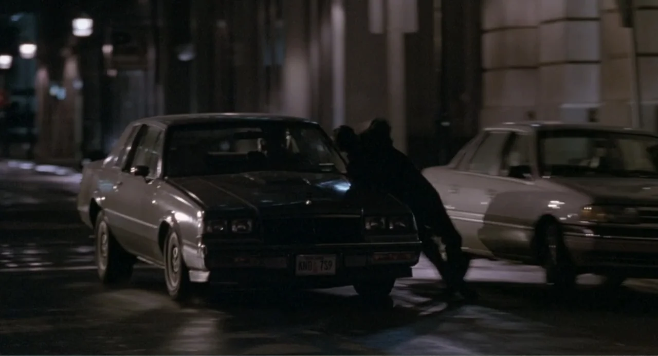 F/X2 (1991) movie still: medium — A dark city street at night features two cars parked on a wet road. A man in a dark coat stands against the front…