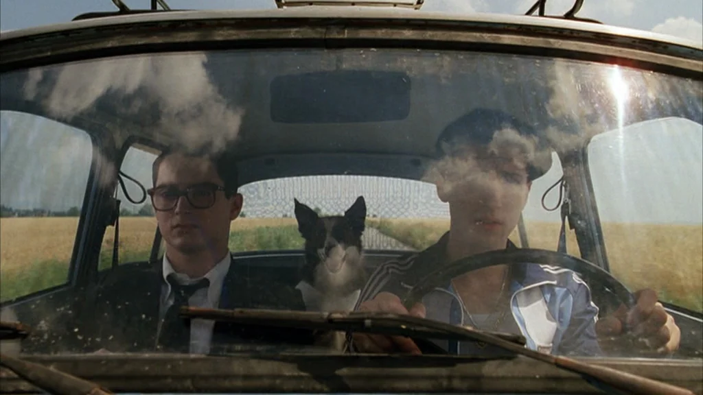 Everything Is Illuminated (2005) movie still: medium — Two men and a dog travel inside a compact car through a rural field. The windshield shows…