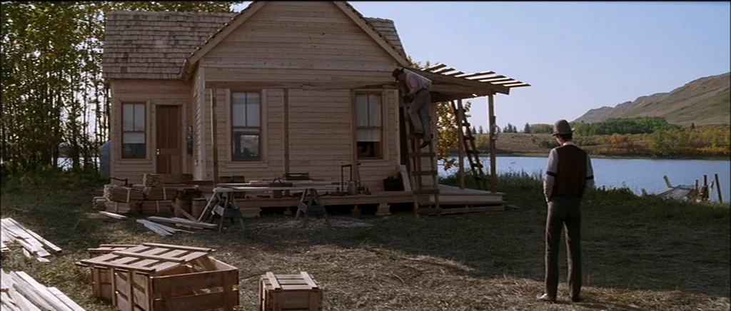 Unforgiven (1992) movie still: wide — An unfinished wooden house stands on a grassy lakeshore next to piles of lumber. One man stands watching while…