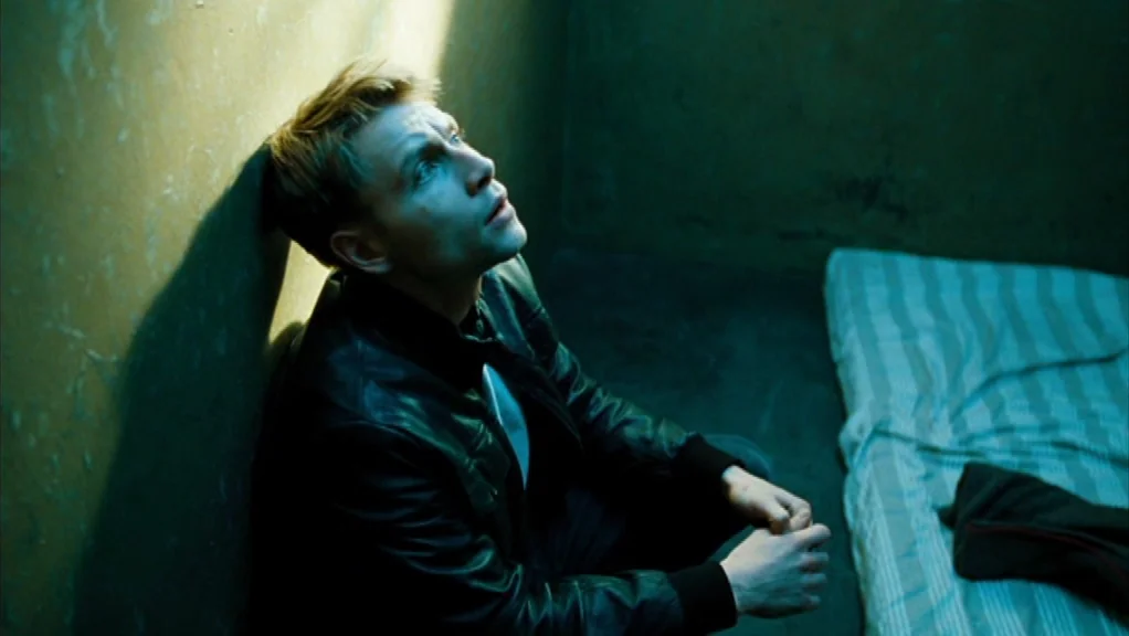 We Are The Night (2010) movie still: medium — A dimly lit room features a worn cot with a striped mattress. A man sits against the wall looking…
