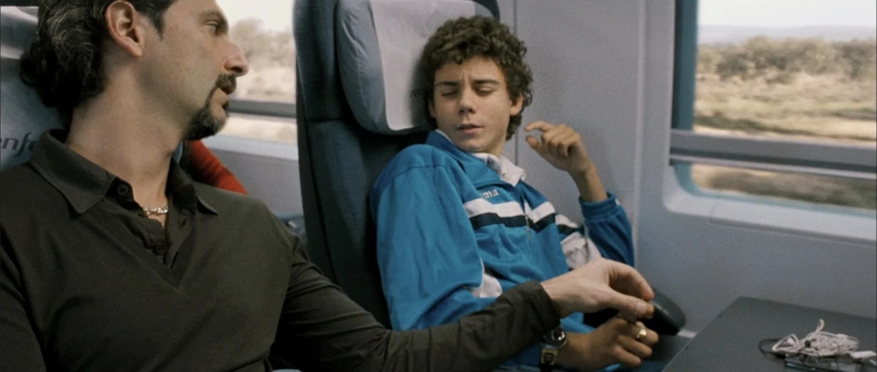 Rivals (1972) movie still: medium — Two individuals sit together in the passenger cabin of a modern rail vehicle. An adult man looks toward a…