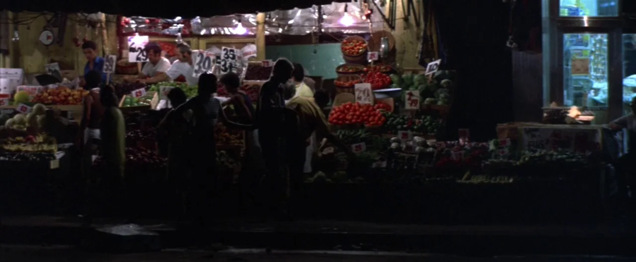 Klute (1971) movie still: wide — A bustling outdoor produce stand is illuminated at night with stacked bins of fruits. Customers in shadow select…