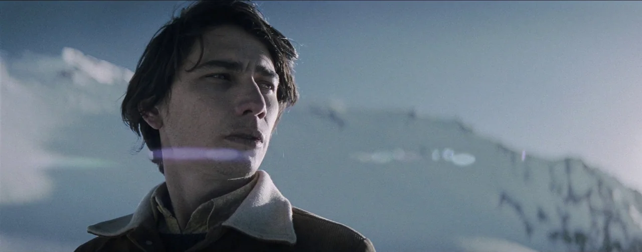 Society of the Snow (2023) movie still: closeup — A snowy mountain landscape serves as a backdrop to a man's face. The man turns his head while…