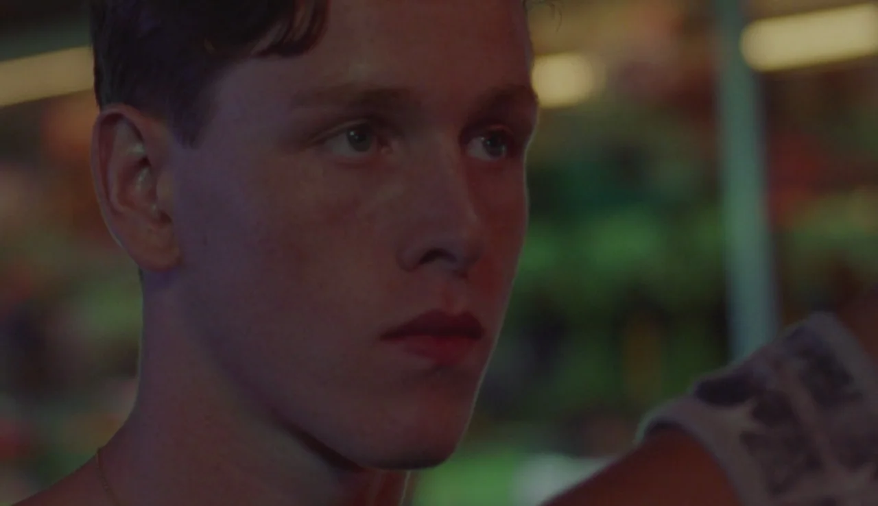 Beach Rats (2017) movie still: closeup — An indoor space is dimly lit by colorful neon signs in the background. A young man looks off-camera while…