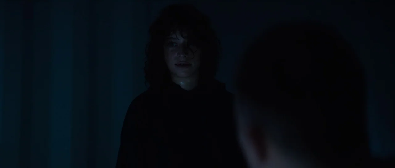 Hellraiser (2022) movie still: medium — A dimly lit room is consumed by deep shadow with very low visibility. A young woman with curly hair faces…