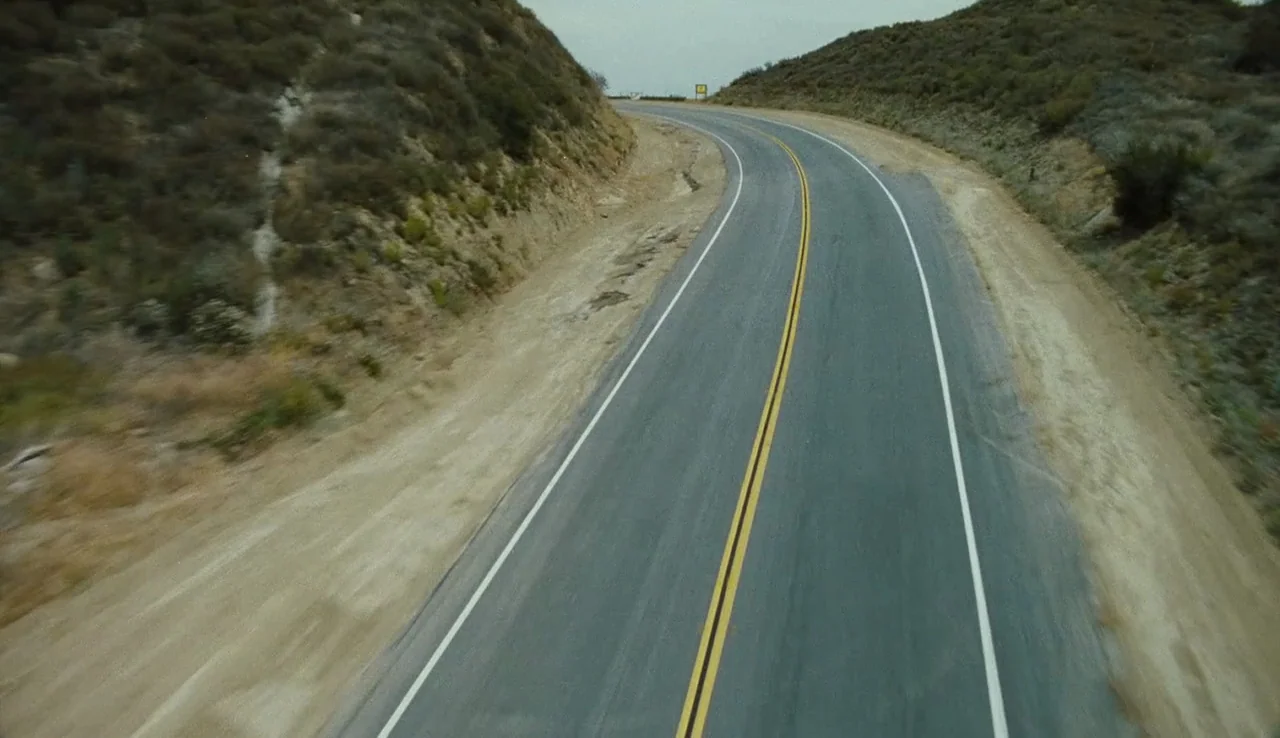 The Devil’s Rejects (2005) movie still: establishing — A rural highway winds through arid, scrub-covered hills under a grey overcast sky. The road…