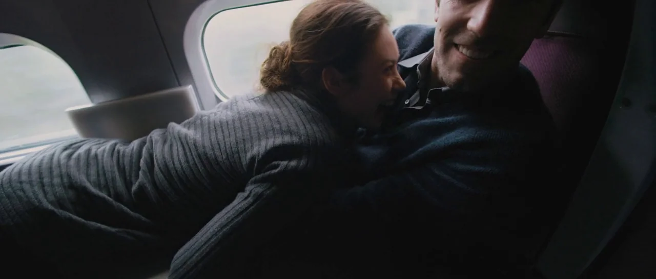 To The Wonder (2012) movie still: medium — Two individuals sit together in a cramped aircraft cabin near an oval window. The woman hugs the man's…
