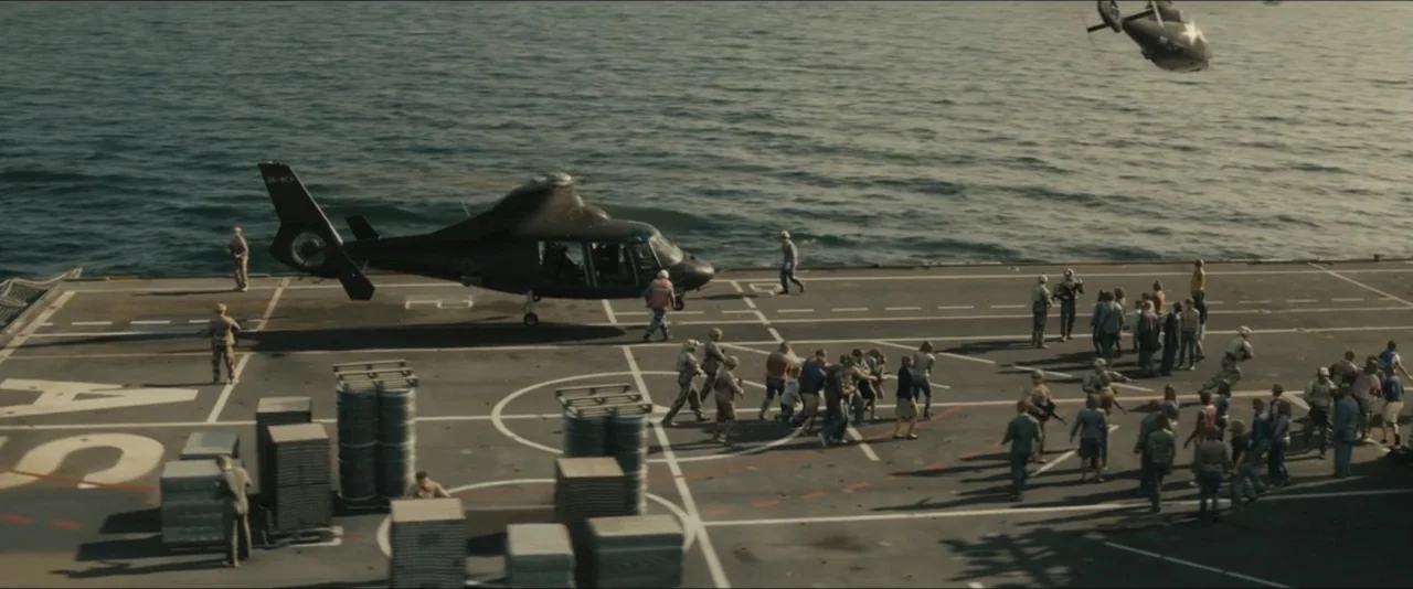 World War Z (2013) movie still: wide — The flat grey surface of an aircraft carrier flight deck sits above dark water. Numerous civilians and…