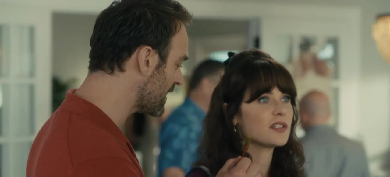 Merv (2025) movie still: medium — A light-filled residential interior features blurred figures in the background. A man stands beside a woman who…