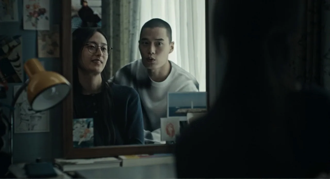 Winter in Sokcho (2025) movie still: medium — A bedroom contains a desk with a mirror and various photos pinned to the wall. A man and a woman stand…