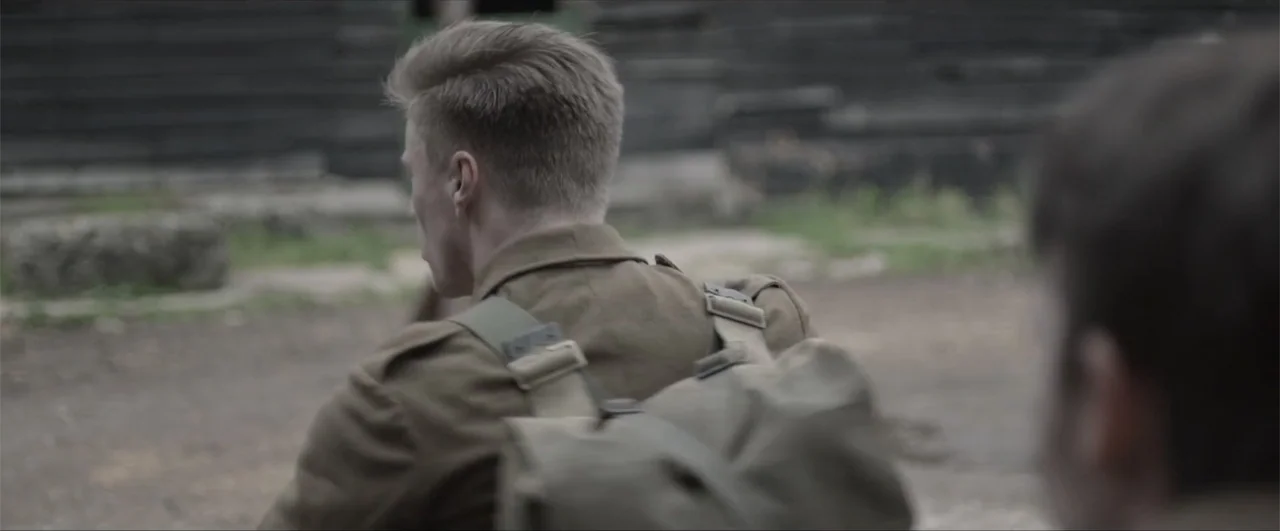 Behind the Line: Escape to Dunkirk (2020) movie still: medium — An outdoor dirt clearing with dark wooden structures in the blurred background. A…