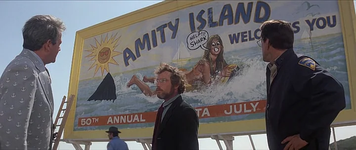 Jaws movie still: wide — A large outdoor billboard displaying a beach scene sits against a clear blue sky. Three men stand together in the…