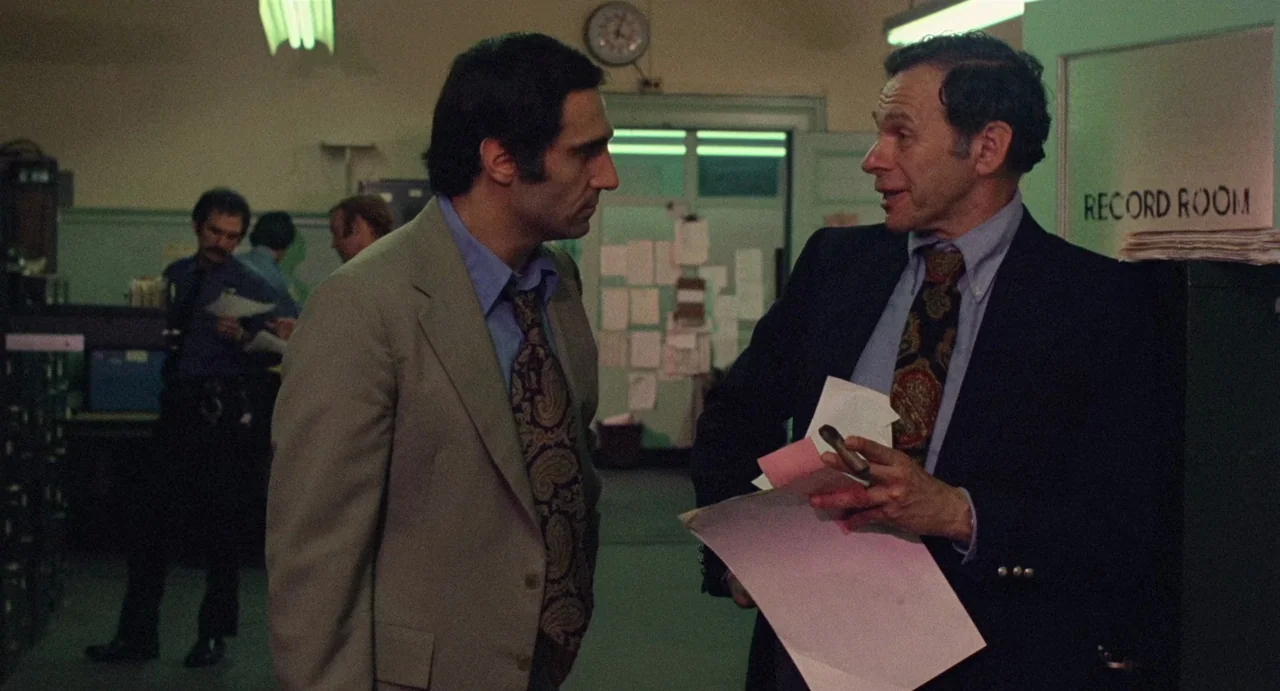 God Told Me To (1976) movie still: medium — A utilitarian room features green-painted walls and a door marked Record Room. Two men in suits stand in…