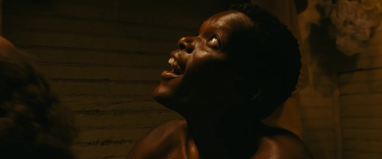 All Dirt Roads Taste of Salt (2023) movie still: closeup — An interior space with horizontal wooden paneling visible behind the subject. A shirtless…