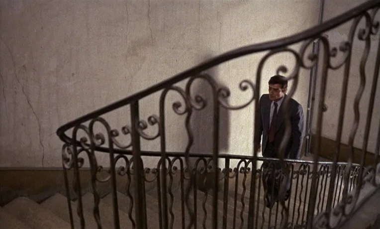 The Man Who Loved Women (1977) movie still: medium — A stone staircase features an ornate, dark wrought-iron railing that curves downward. A man in…