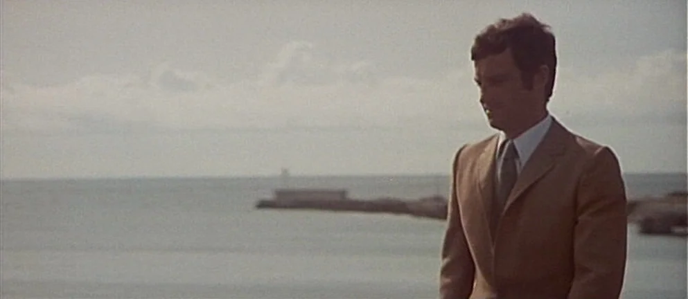 Mississippi Mermaid (1969) movie still: medium — A man stands along a coastal shoreline on an overcast day. He looks toward the horizon near a stone…