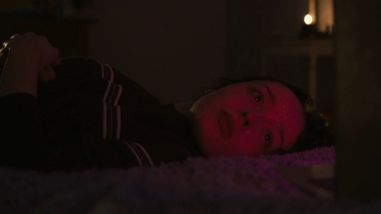 We’re All Going to the World’s Fair (2021) movie still: closeup — A dark bedroom with the subject lying on a textured gray carpet floor. The…