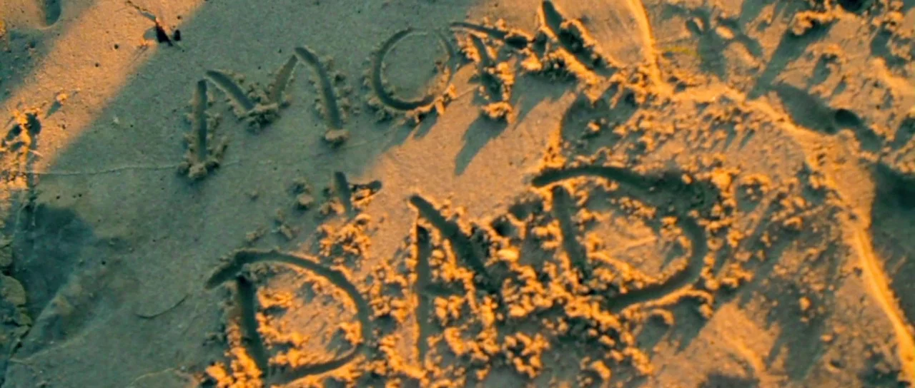 I Melt With You (2011) movie still: closeup — The word MOM is inscribed above the word DAD in moist, textured sand. Strong sunlight highlights the…