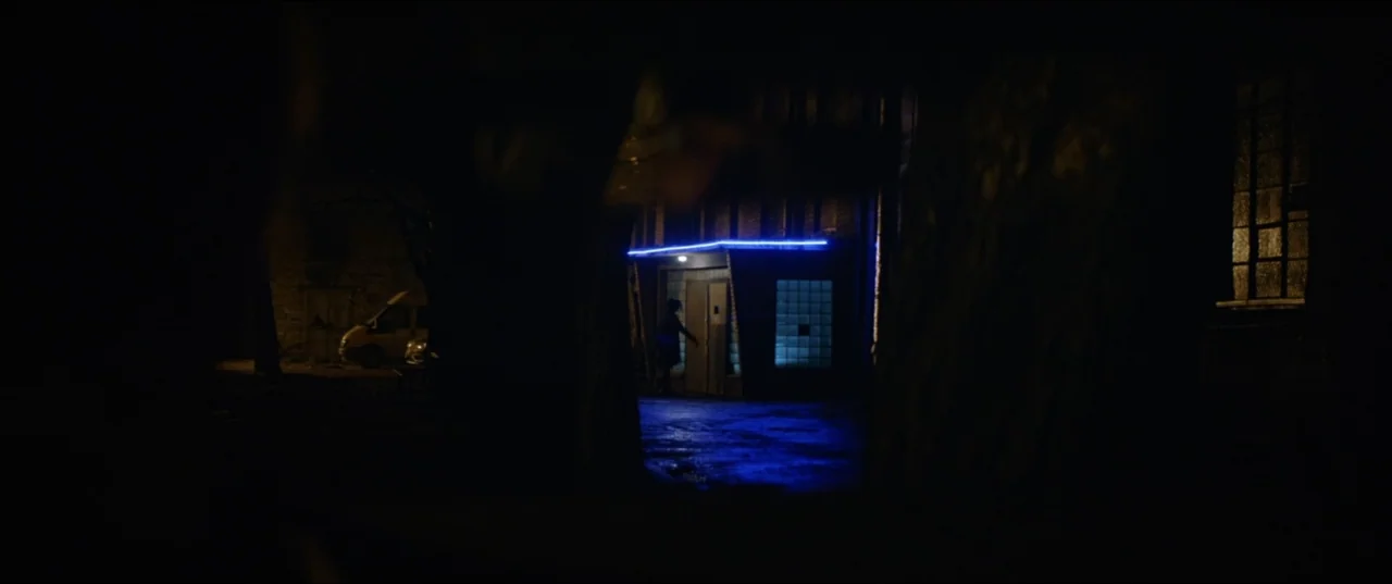 Dogs Don't Wear Pants (2019) movie still: wide — A dark outdoor space features a building entrance illuminated by blue neon lighting. Trees frame…