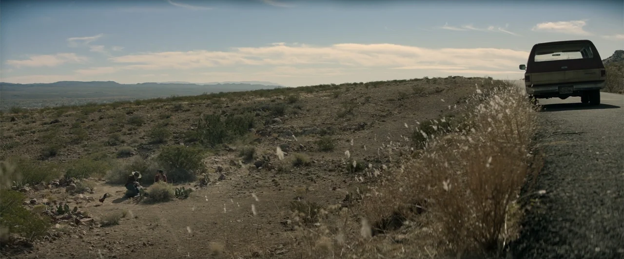 Cry Macho (2021) movie still: wide — A vast, dry desert landscape stretches beneath a clear blue sky. Two people crouch near a parked car while the…