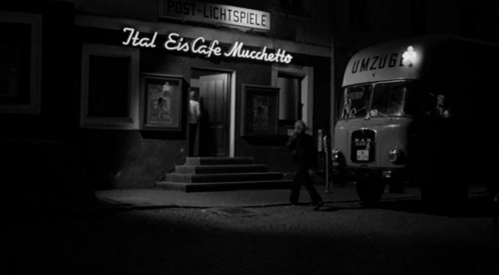 Kings of the Road (Im Lauf der Zeit) (1976) movie still: wide — A nighttime street scene features a building with a brightly lit neon sign. A person…